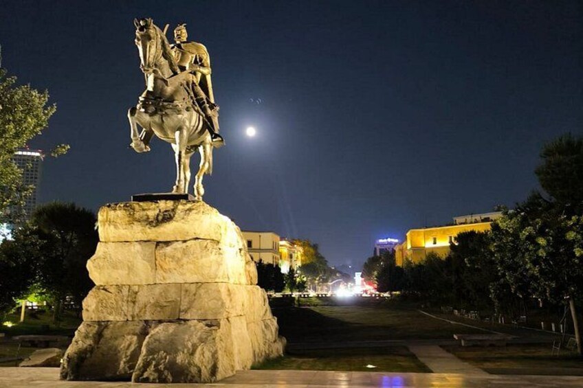 A statue of a man on a horse, a stone pedestal, and a cityscape with buildings and a street at night. 1
