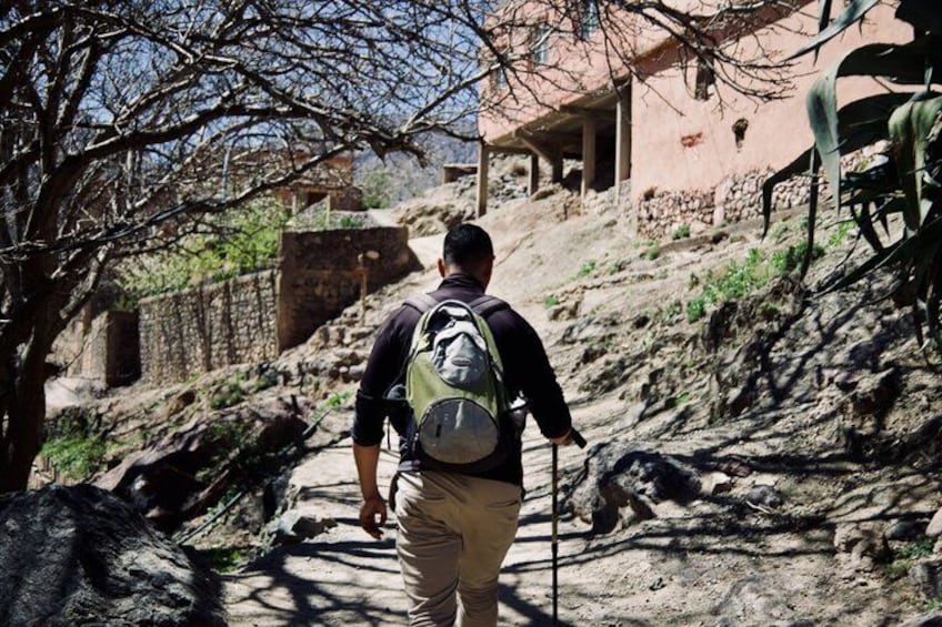 A person hiking on a rocky trail with a backpack, surrounded by trees and a building in the background. 3