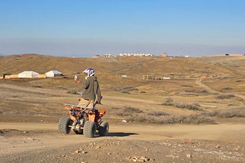 Una persona guida un ATV su una strada sterrata in zona rurale, con vegetazione rada e alcuni edifici in lontananza. 4