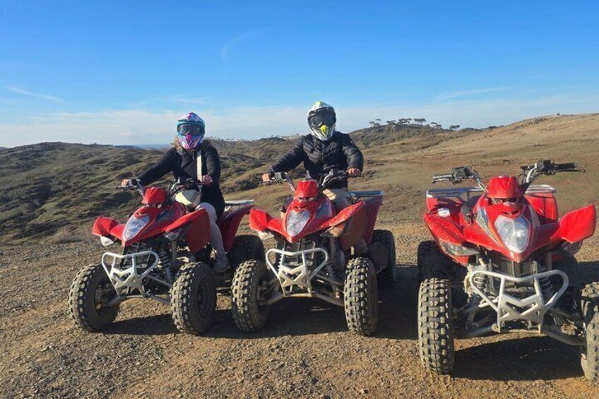 Tre persone su quad rossi su una strada sterrata, con un paesaggio collinare sullo sfondo. 1