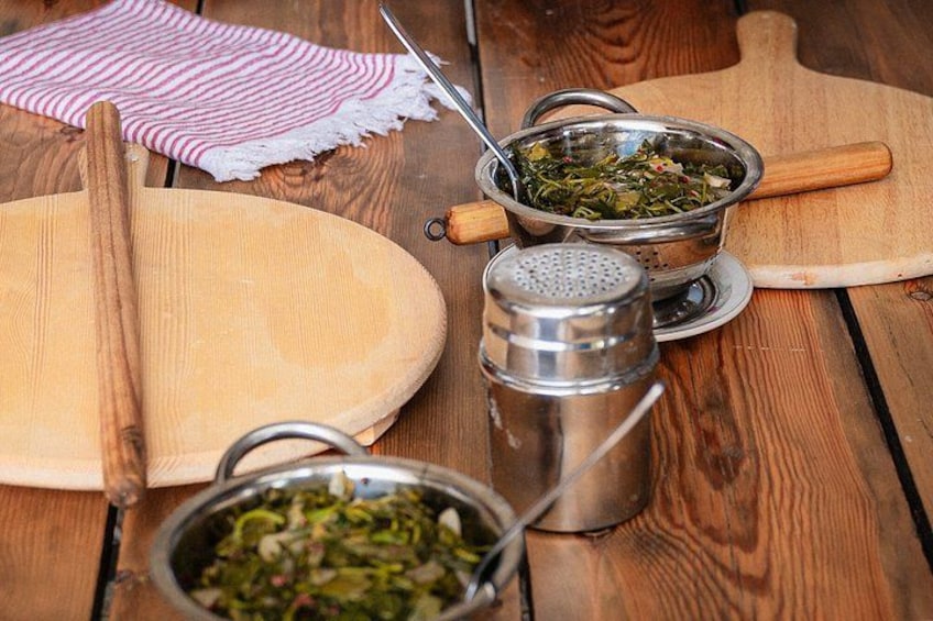 A wooden table with a rolling pin, a metal colander with greens, a salt shaker, and a wooden cutting board. 2