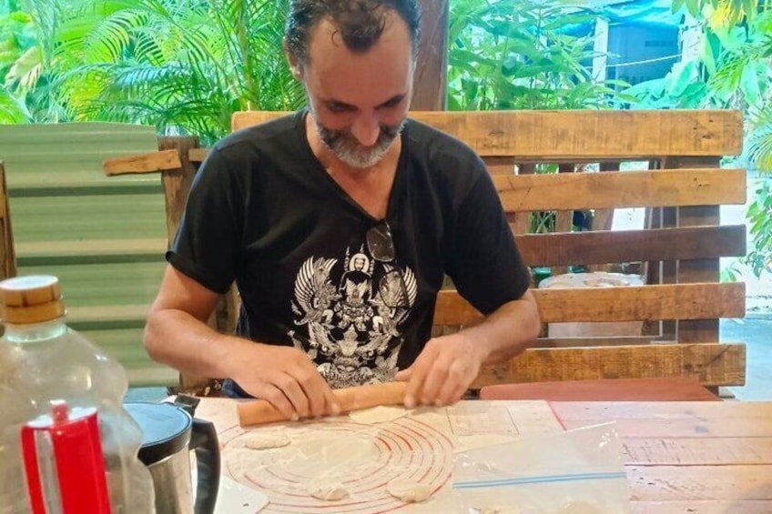 A person rolling dough on a wooden table with a rolling pin, surrounded by green plants. 4