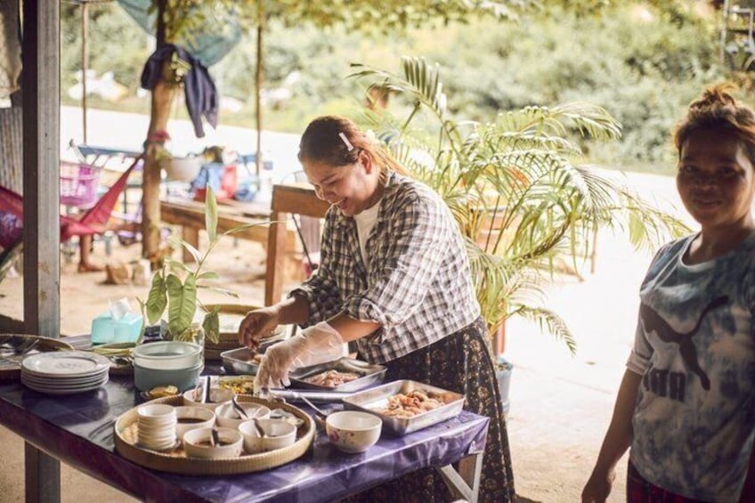 An outdoor dining area with a person serving food, a table with dishes, and a tropical setting with plants. 1