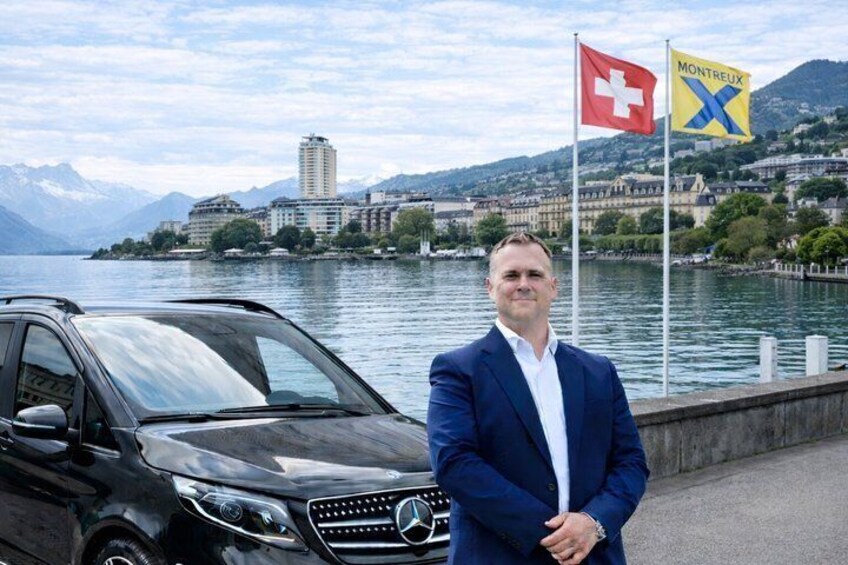 A man in a blue suit standing next to a black Mercedes-Benz SUV with flags of Switzerland and Montreux in the background. 1