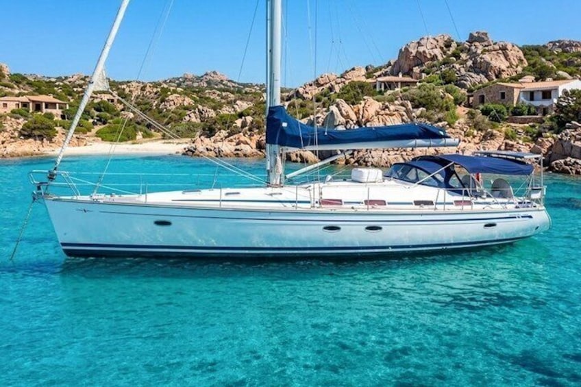 A sailboat on turquoise water with rocky hills and houses in the background. 1