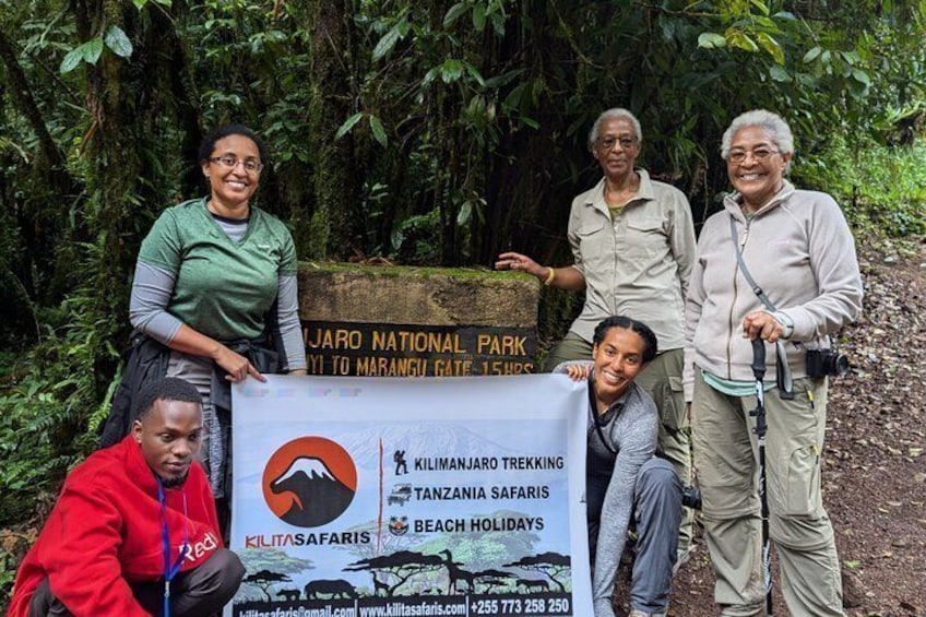 Eine Gruppe von Menschen steht vor einem Schild für Kilimanjaro-Besteigungen, Tansania-Safaris und Strandurlaub. 2