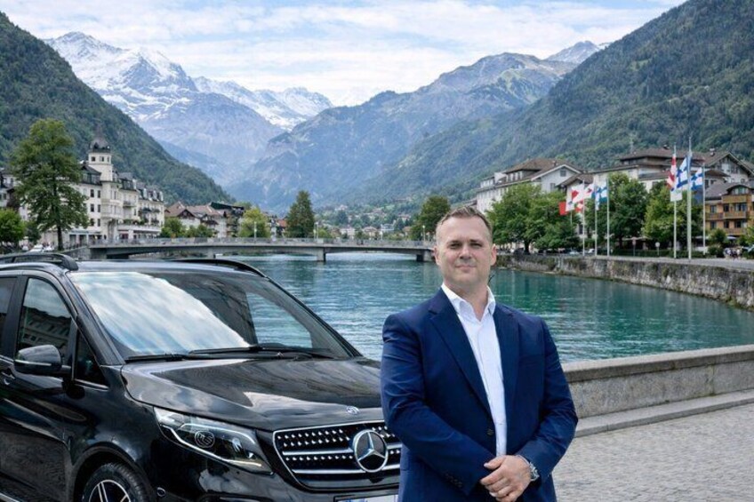 En man i en blå kostym står bredvid en svart Mercedes-Benz-SUV i en stad vid en sjö, med berg i bakgrunden. 1