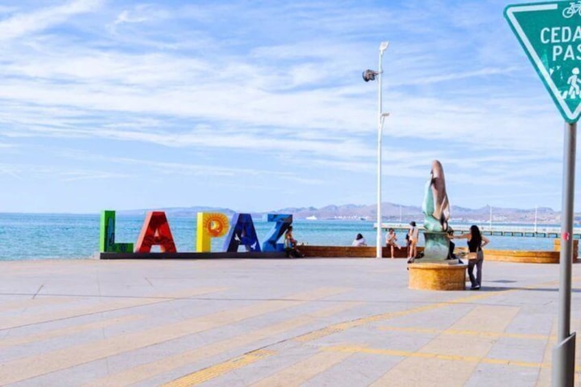 Un letrero colorido que dice 'LAPAZ', una estatua y una señal de 'CEDA PAS' junto a la playa. 5