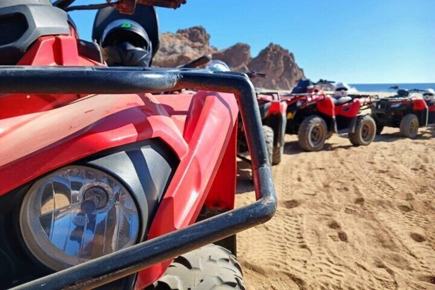 Een rode quad op een zandstrand, met andere quads op de achtergrond en een rotsformatie. 1