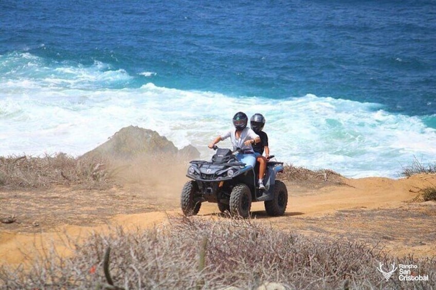 Twee personen op een quad op een zanderig strand, met oceaan golven op de achtergrond. 5