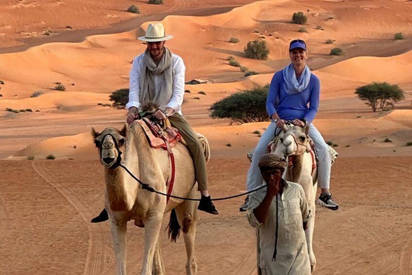 Two people riding camels in a desert landscape with sand dunes. 2