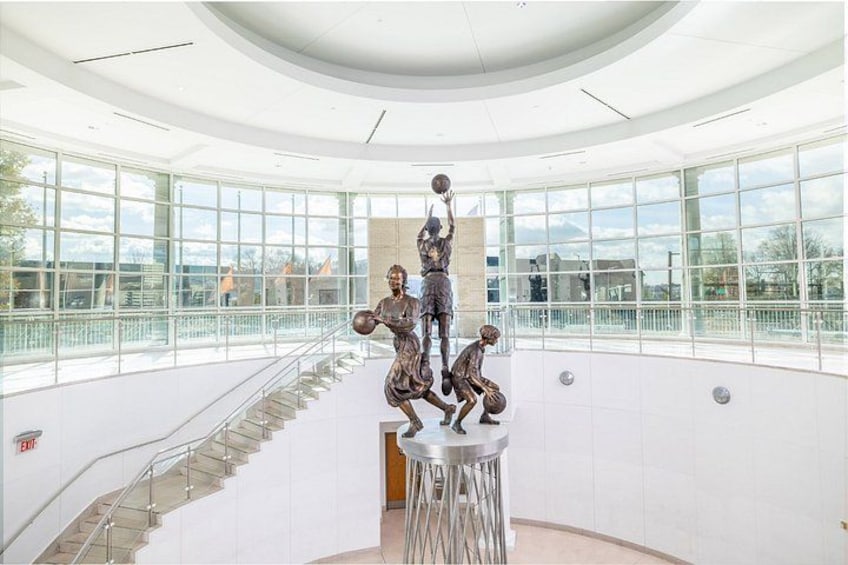 A modern building interior with a central staircase, large windows, and a sculpture of basketball players. 2