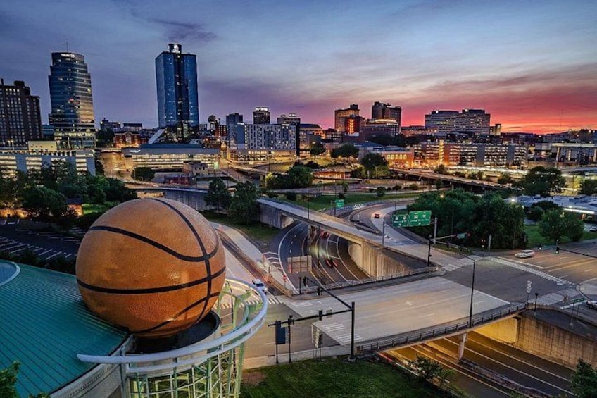 A cityscape at dusk with a large basketball in the foreground, a highway interchange, and high-rise buildings. 1