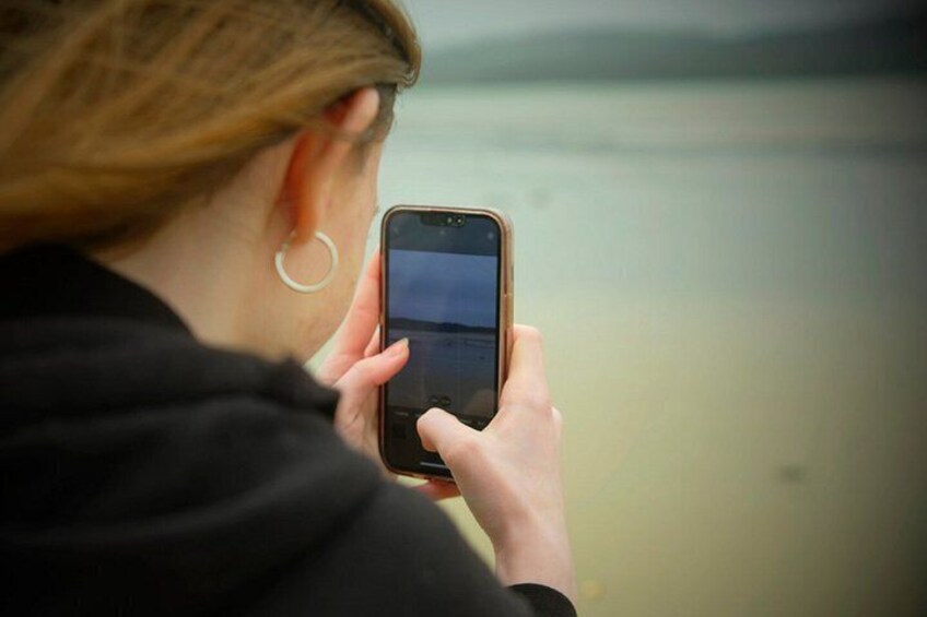 A person holding a smartphone, facing away from the camera, with a body of water in the background. 4