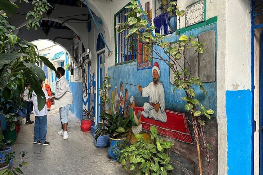 Un stretto vicolo con un murale raffigurante una figura seduta, piante in vaso e la facciata di un edificio blu e bianco. 2