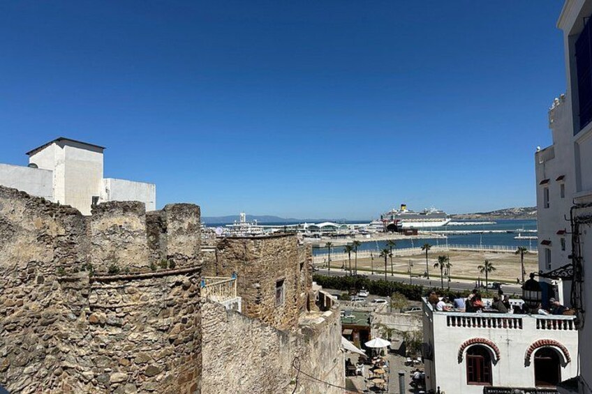 Un paisaje costero con murallas de piedra históricas, un edificio blanco con arcos y un moderno crucero amarrado en el puerto. 3