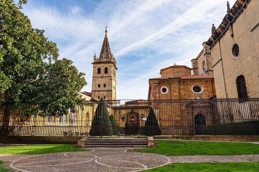 Un edificio histórico con una torre, un patio empedrado con una fuente y un camino bordeado de árboles. 5