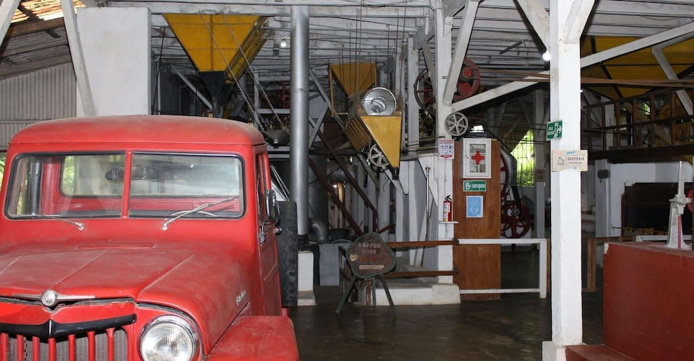 A red vintage car in a garage with industrial machinery and a sign indicating a coffee tour. 2