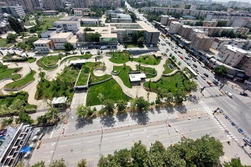 An urban area with modern buildings, a central green space with pathways, and a busy road. 2