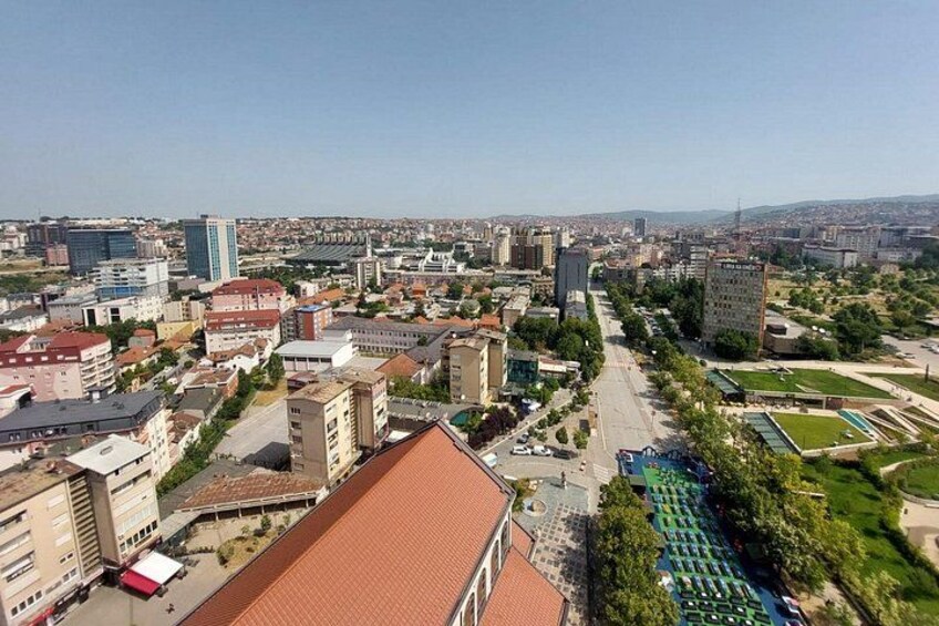 A cityscape with buildings, a clear sky, and a road with greenery on the side. 2