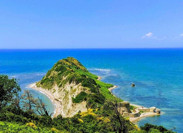 A coastal landscape with a prominent green hill, clear blue sea, and rocky shoreline. 2