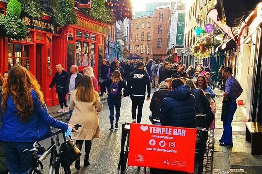 A busy street with people walking, a red building labeled 'TEMPLE BAR,' and a signboard with the bar's name and website. 1