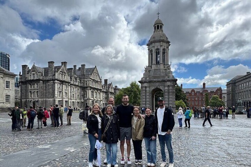 A group of people standing in a cobblestone square with historic buildings and a clock tower in the background. 1