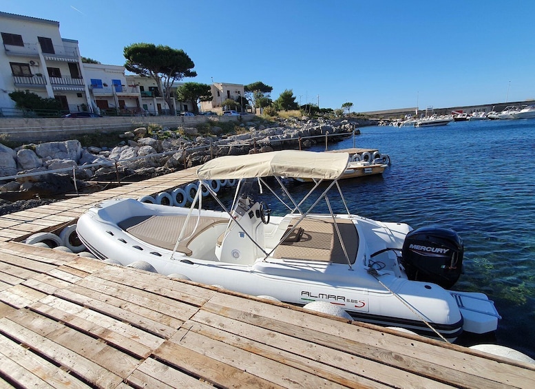 A white motorboat named 'ALMAR 585' docked at a wooden pier with a rocky shoreline and buildings in the background. 5