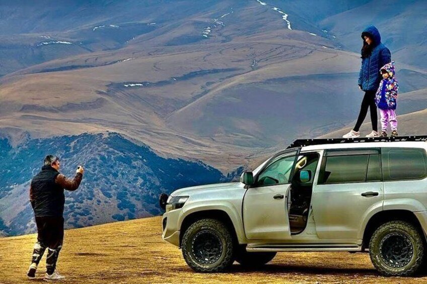 Una persona fotografiando con una cámara, un niño de pie sobre un vehículo y un paisaje montañoso al fondo. 4