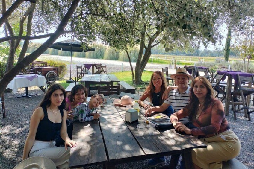 A group of people sitting at a wooden table outdoors, with trees and a grassy field in the background. 3