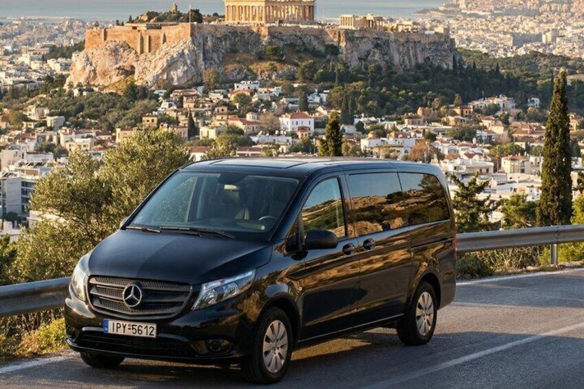 En sort Mercedes-Benz varevogn parkeret på en vej med udsigt til en by og oldtidsruiner i baggrunden. 2