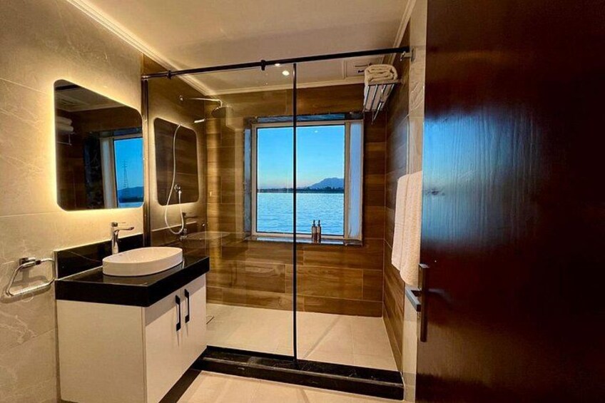 A modern bathroom with a glass shower enclosure, a sink, and a large window overlooking a body of water. 1