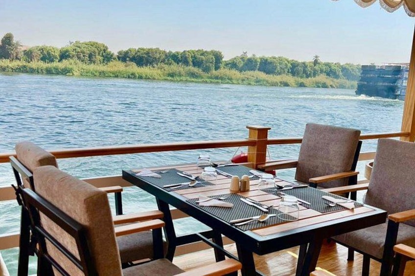 A dining table set on a boat with chairs, overlooking a river with greenery and a distant ship. 4