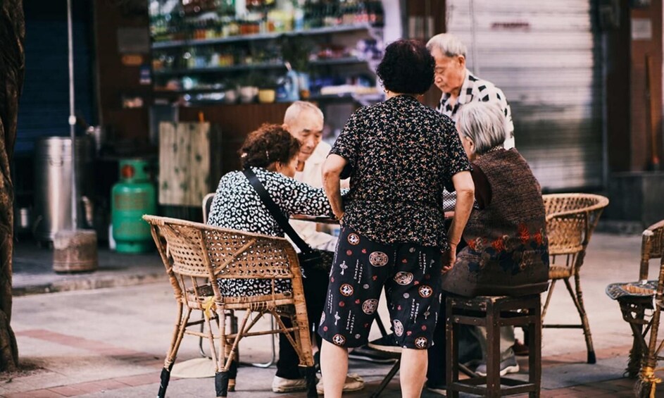 A group of people sitting at wicker chairs on a sidewalk, with a store in the background. 2
