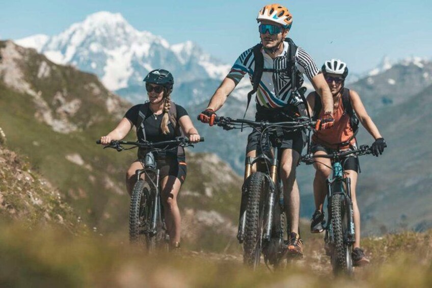 Trois personnes font du vélo tout terrain sur un sentier, avec des montagnes enneigées en arrière-plan. 1