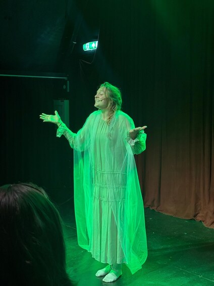 A person in a flowing white dress standing on a stage with arms outstretched, under green lighting. 4