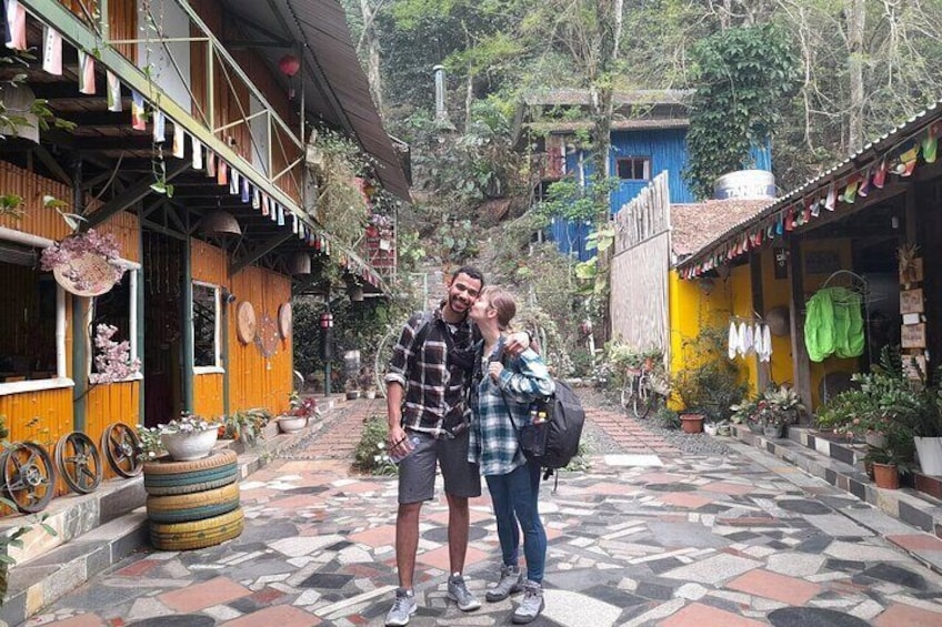 A couple standing in a colorful courtyard with wooden buildings, potted plants, and a patterned tile floor. 1
