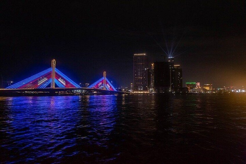 A brightly lit bridge with two towers, a body of water in the foreground, and city buildings in the background. 3