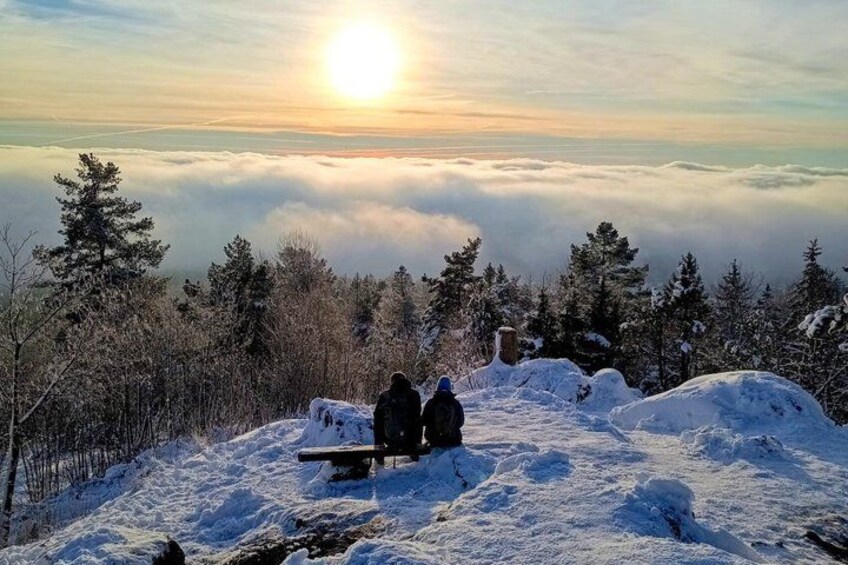 A snowy landscape with a bench and two people sitting, overlooking a forest and a sunlit horizon. 2
