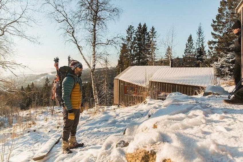 A person standing on a snow-covered slope with a backpack, a wooden structure in the background, and a forested area. 3