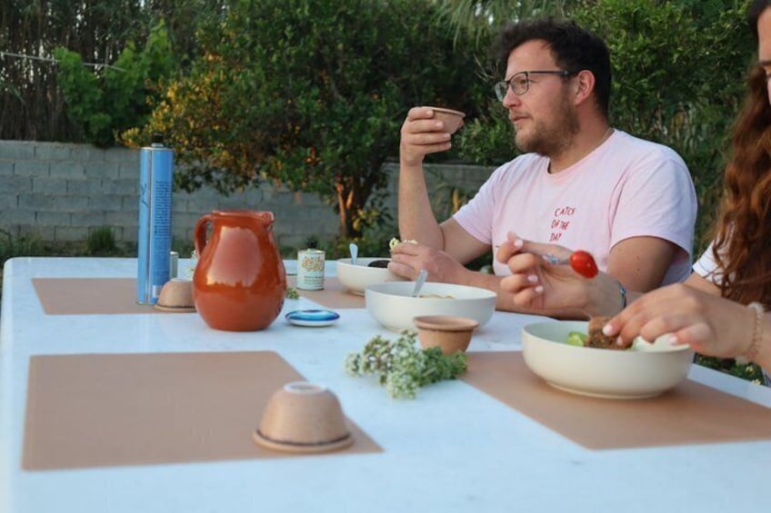 Een groep mensen geniet van een maaltijd buiten aan een tafel, met verschillende gerechten en een karaf. 1