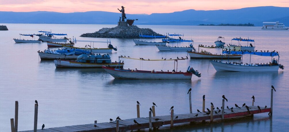 A calm lake with several boats docked, a statue on a small island, and birds perched on the dock. 4