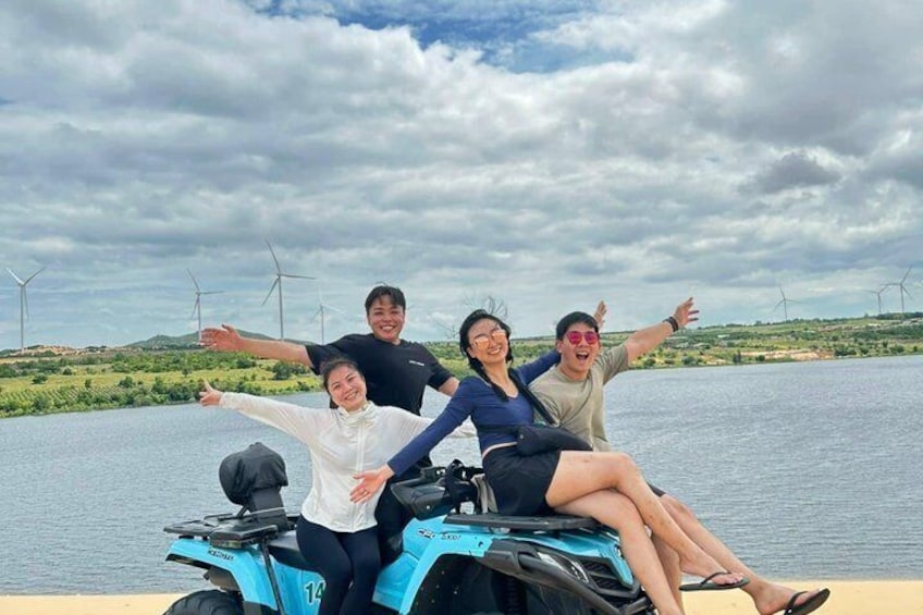 Four people on a blue ATV with a lake and wind turbines in the background. 2