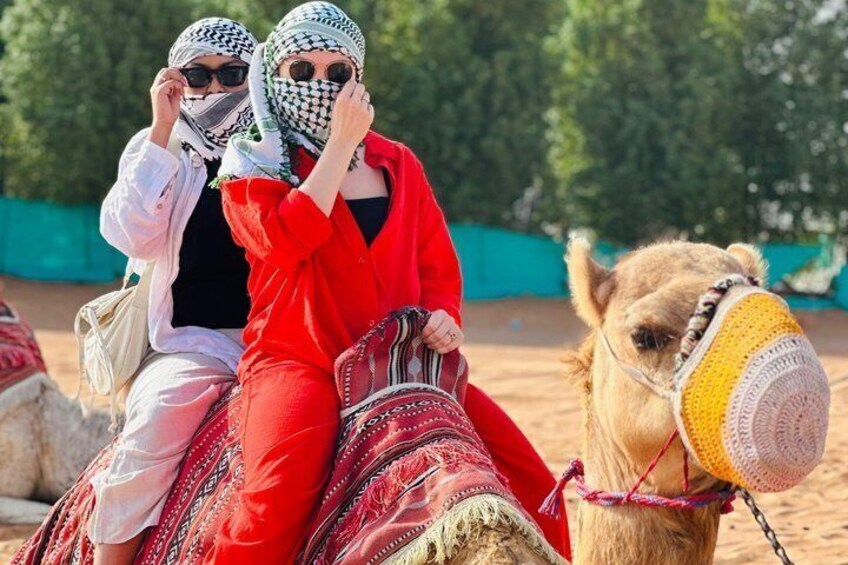 Two people riding a camel with a decorated saddle, in a desert-like environment. 2
