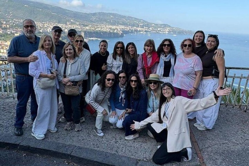 A group of people posing on a coastal overlook with a view of the sea and distant cityscape. 3