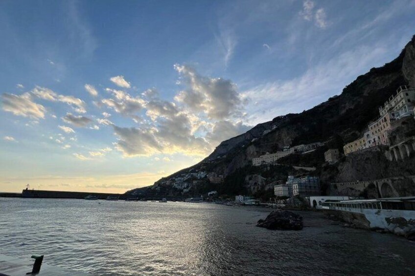 A coastal town with buildings on a hillside, a calm sea, and a clear sky. 1