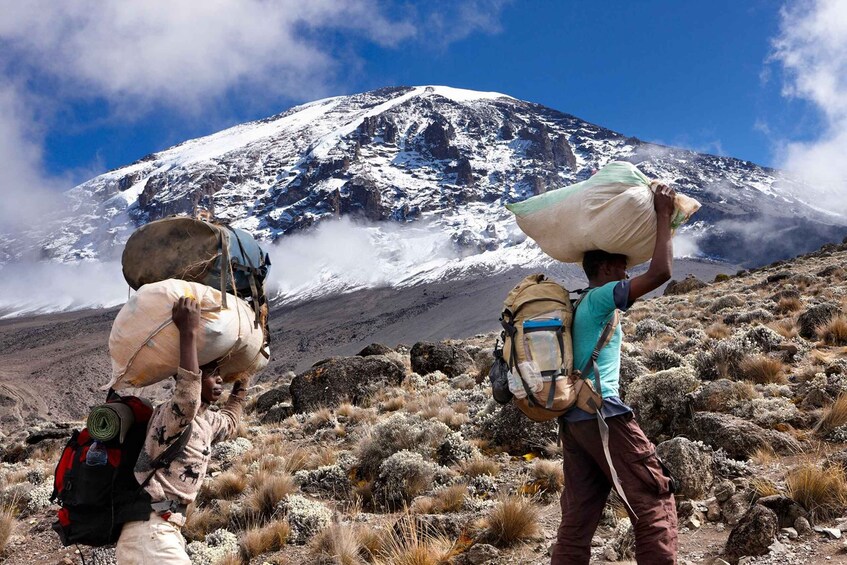 Two hikers carrying large bags across a rocky terrain with a snow-capped mountain in the background. 2