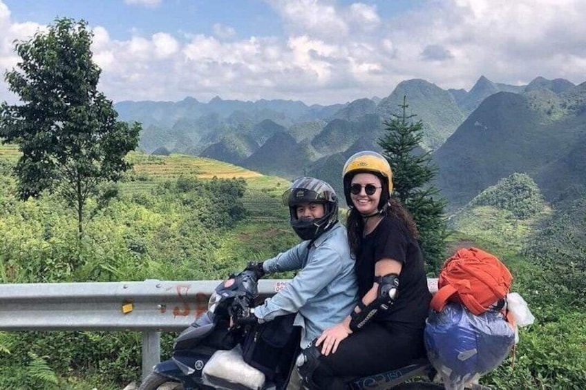 Two people sitting on a motorcycle, surrounded by lush green mountains and a clear sky. 1