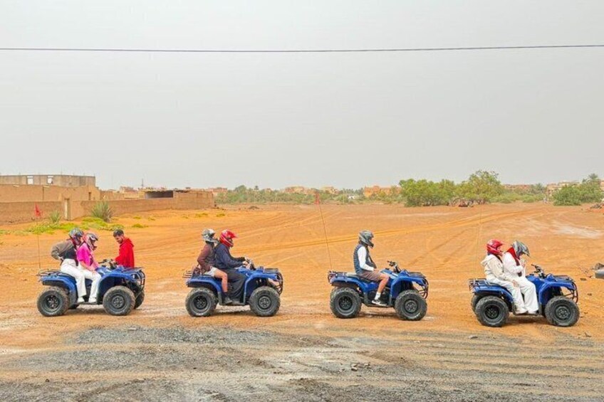 Four people riding blue ATVs on a dirt road with a barren landscape and distant buildings. 3