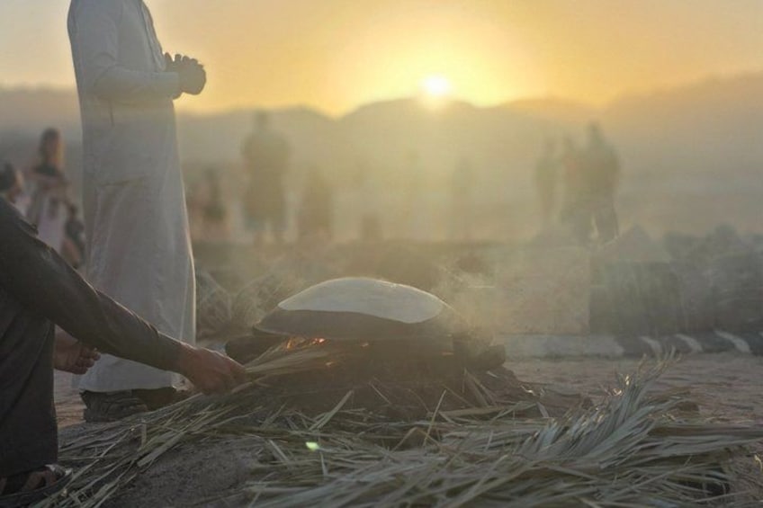 A person cooking over an open fire with a large pot, surrounded by dry grass and a sunset in the background. 2